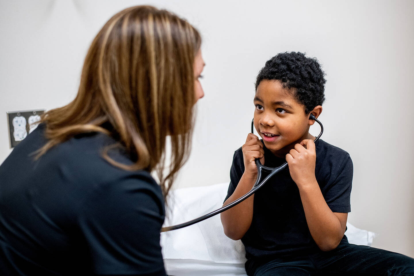A young child holds a stethoscope to their chest while engaging with a healthcare provider in a clinical setting.
